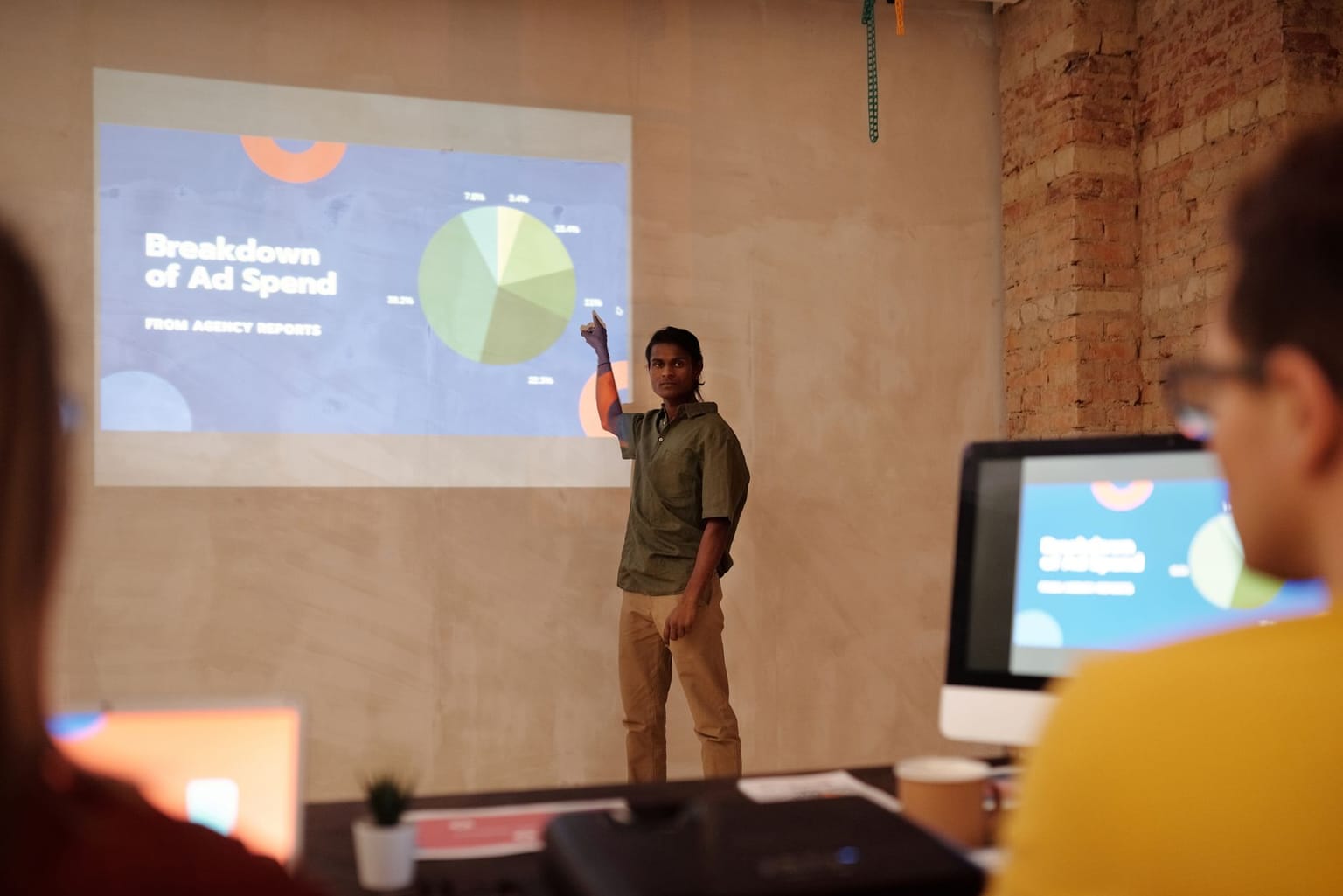 A person stands and points to a projected presentation slide showing a pie chart titled “Breakdown of Ad Spend,” highlighting the impact of effective