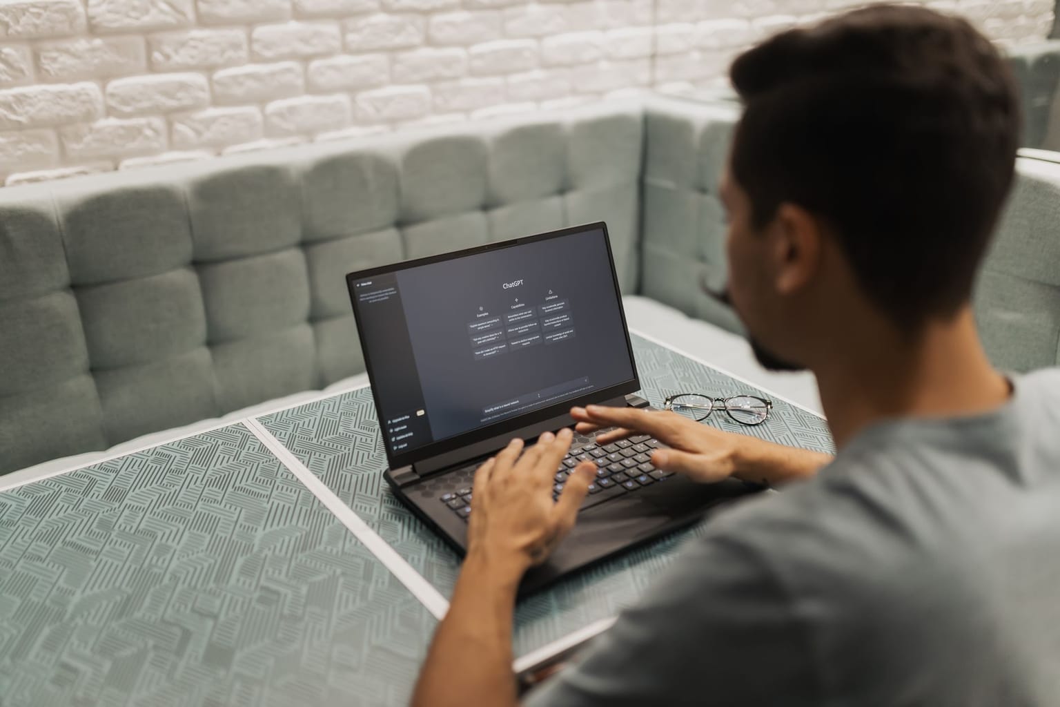 A person types on a laptop at a patterned table in a booth. Glasses rest nearby, and the laptop screen displays an AppSumo AI tools dashboard