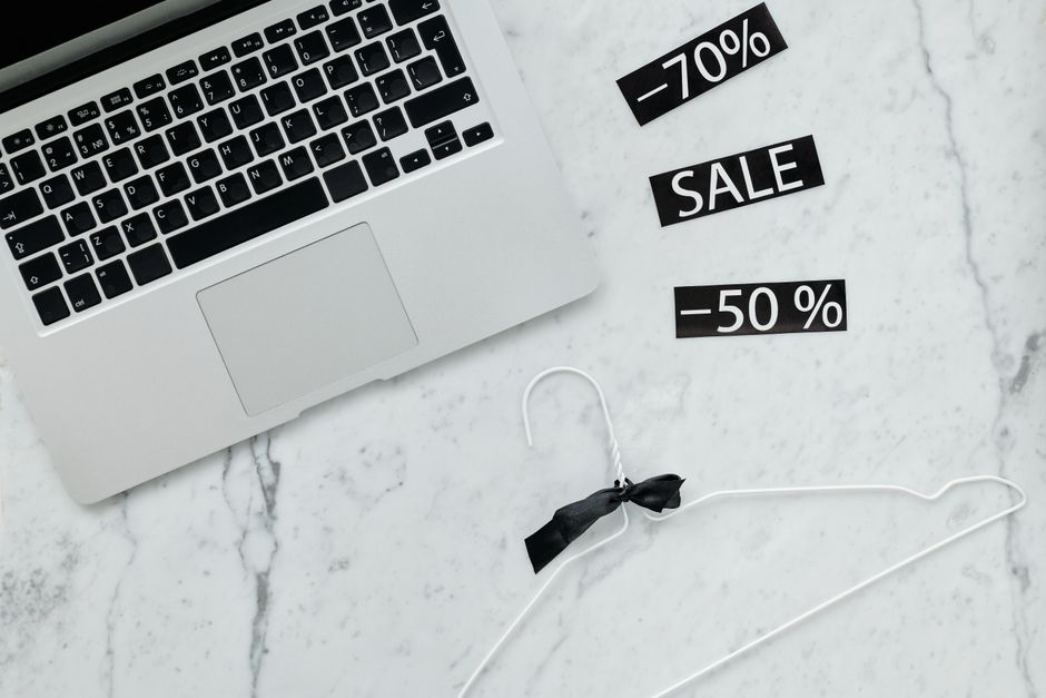 A laptop, sale signs reading "-70%", "SALE" and "-50%", and a white hanger with a black ribbon on a marble surface