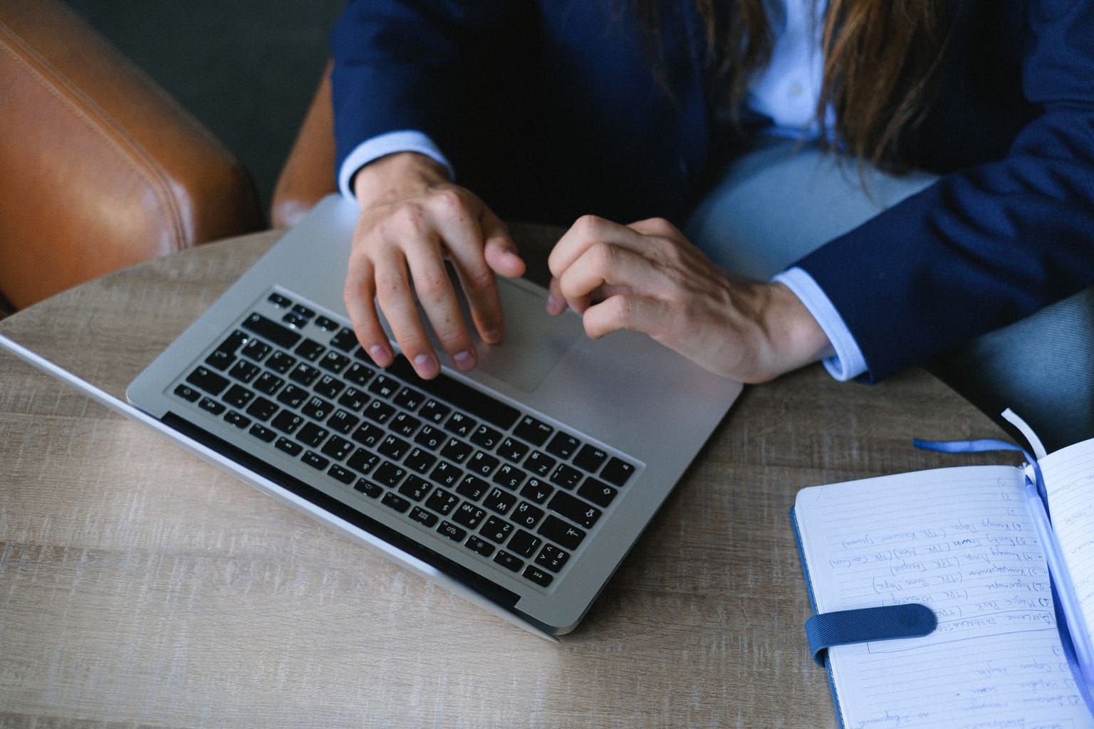 A person in a blue blazer types on a laptop at a wooden table. Next to the laptop is an open notebook with handwritten notes about AppSumo pricing