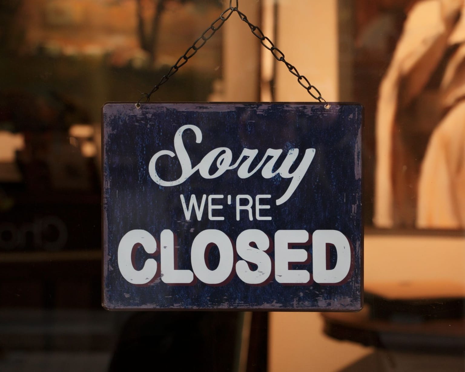 A blue and white sign hanging on a door reads “Sorry We’re Closed,” signaling a closed business. The background is out of focus
