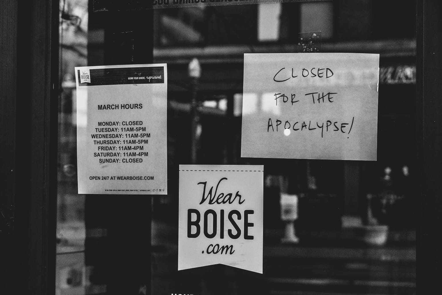 A black and white photo shows a closed business’s window with signs stating hours, a website