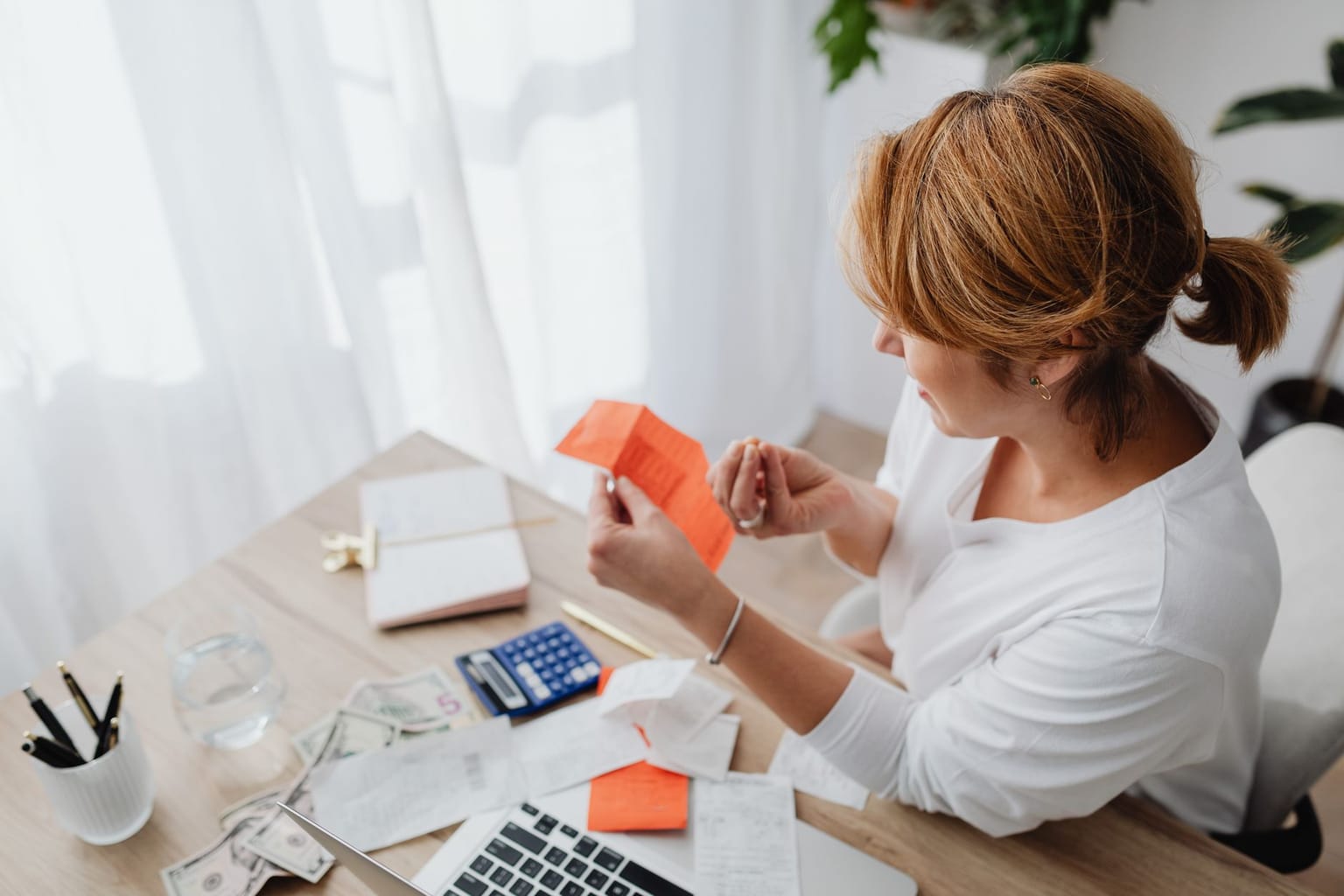 A person reviews a receipt at a desk with a laptop, calculator, notebook and cash