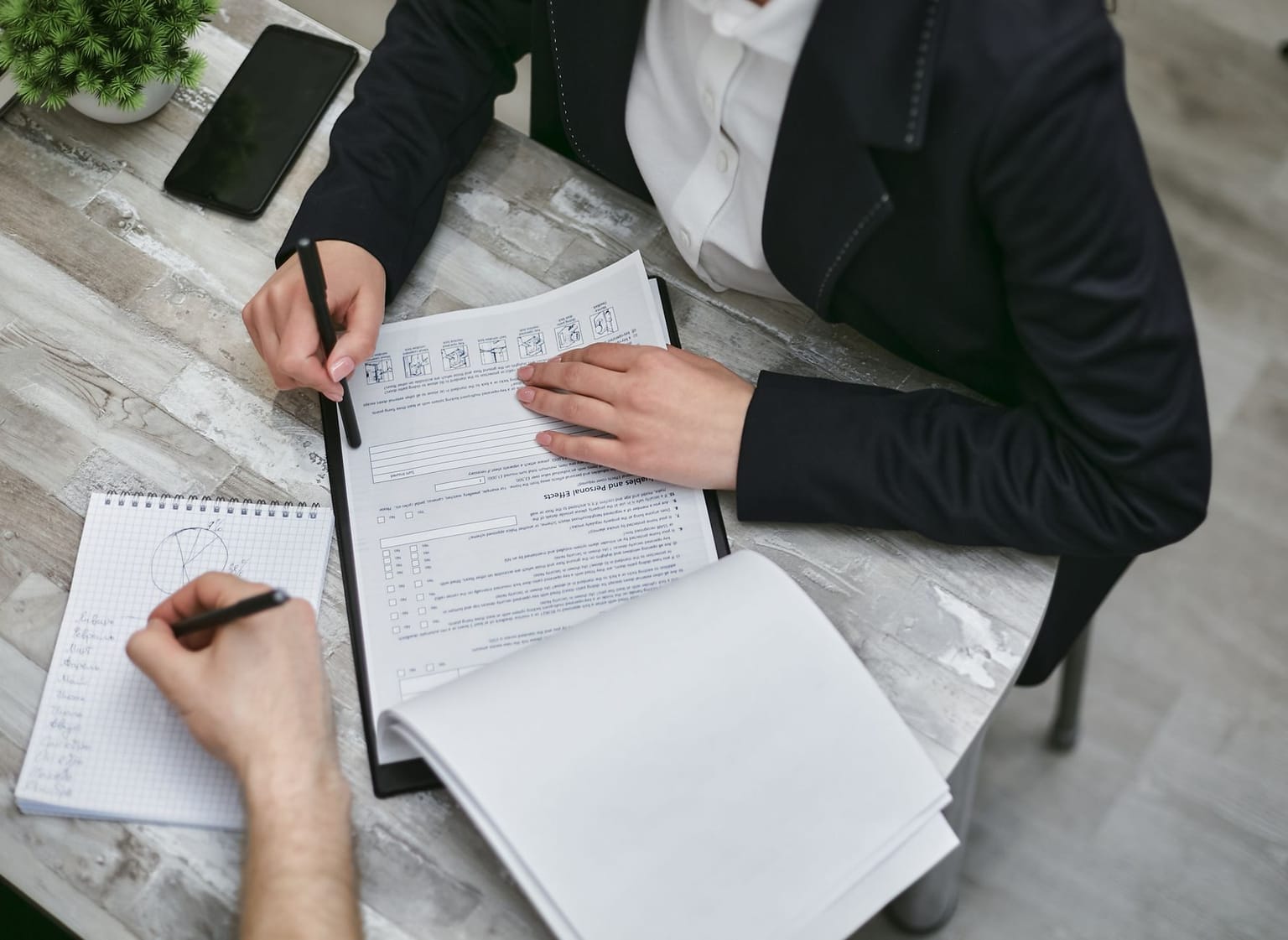 Two people sit at a table filling out paperwork; one is writing on a clipboard while the other takes notes on graph paper