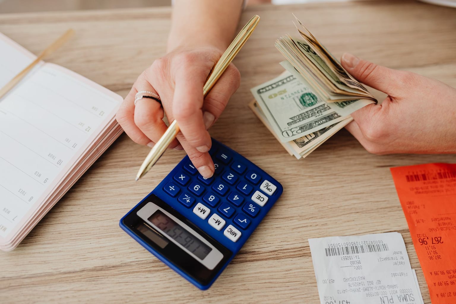 A person holding a stack of U.S. dollar bills in one hand and using a blue calculator with the other