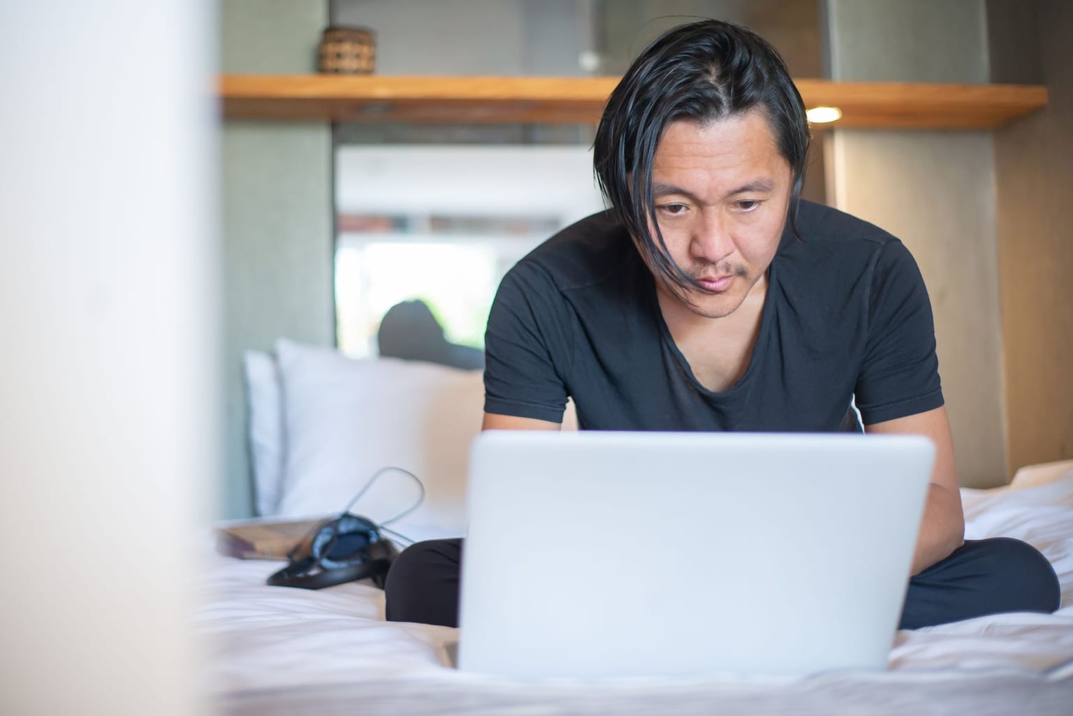 Person with straight dark hair sits cross-legged on a bed, focused on a laptop in front of them. Headphones and a book are beside them