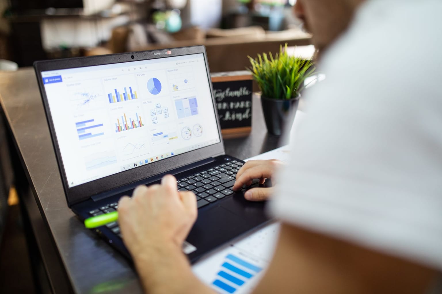 A person works on a laptop displaying charts and graphs from spreadsheet software. The individual holds a green highlighter