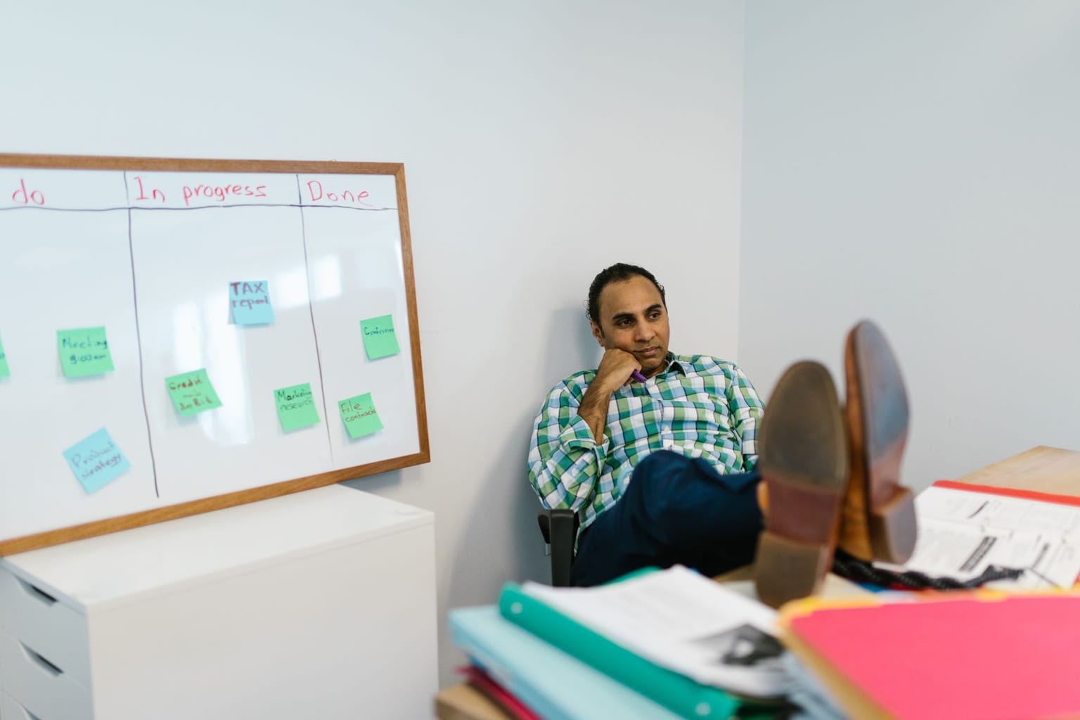 A person sits with feet up on a desk, looking thoughtful. Beside them is a whiteboard displaying “To do,” “In progress,” and “Done” columns with sticky