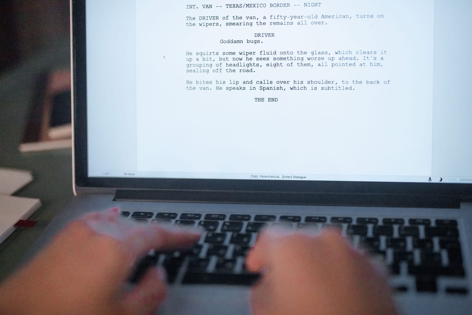 A person types on a laptop, using a grammar checker as the screen displays a movie script mentioning a scene at the Texas/Mexico border, a driver