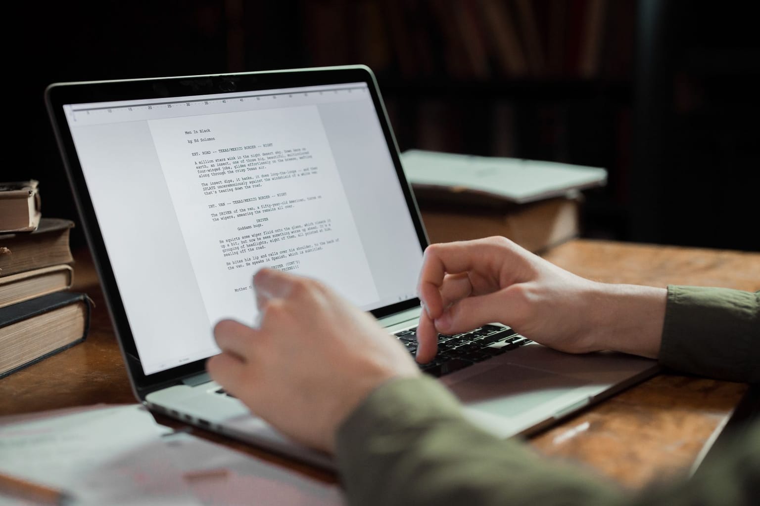 A person types on a laptop at a wooden desk with papers and books around, using a reliable writing tool and grammar checker while working on a document