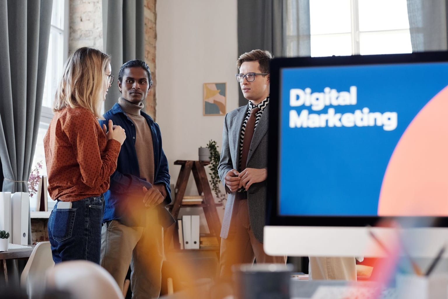 Three people stand and talk in an office with a computer screen in the foreground displaying Digital Marketing, sunlight streaming through windows