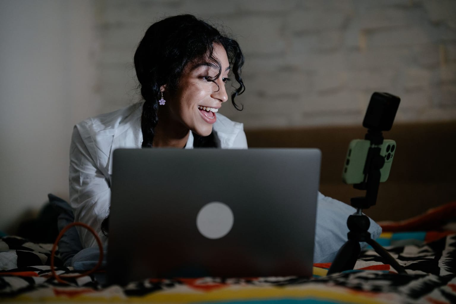 A woman smiles while looking at a laptop and a smartphone on a tripod, possibly video messaging or using a screen recorder