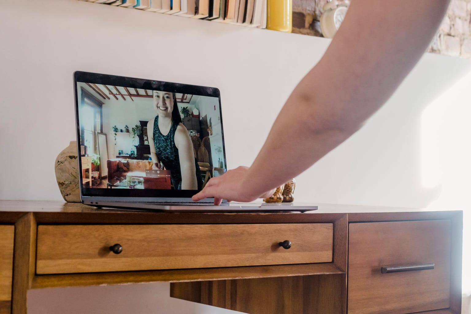 A person’s hand touches a laptop on a wooden desk, with a video call showing another person smiling and wearing a sleeveless shirt. Shelves and books are