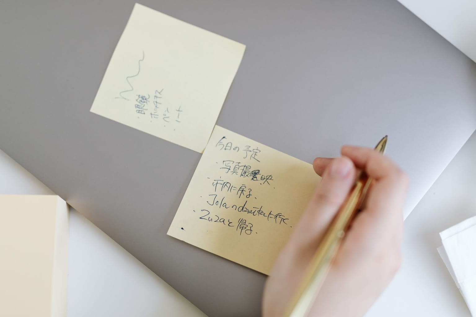 A hand holding a pen above a sticky note with handwritten text in Japanese, placed on a closed silver laptop