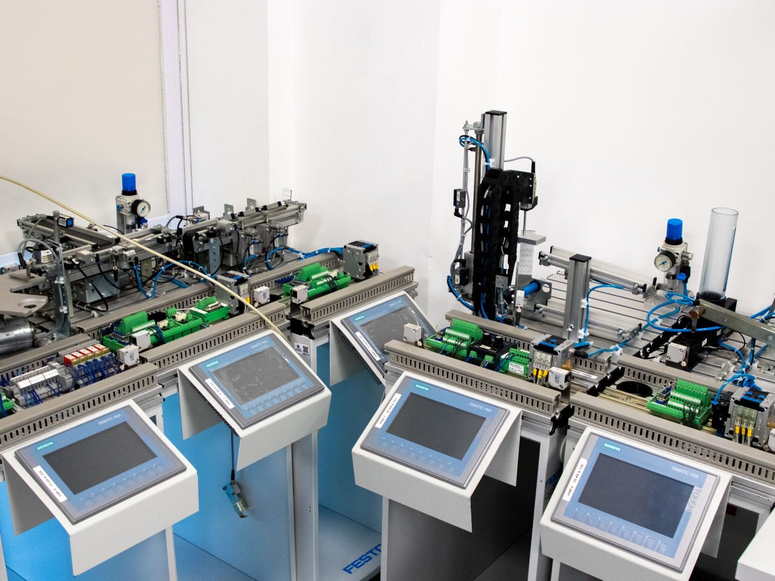 Four industrial control panels with touchscreens and various wiring, tubes, and automation components are arranged in a lab or factory setting