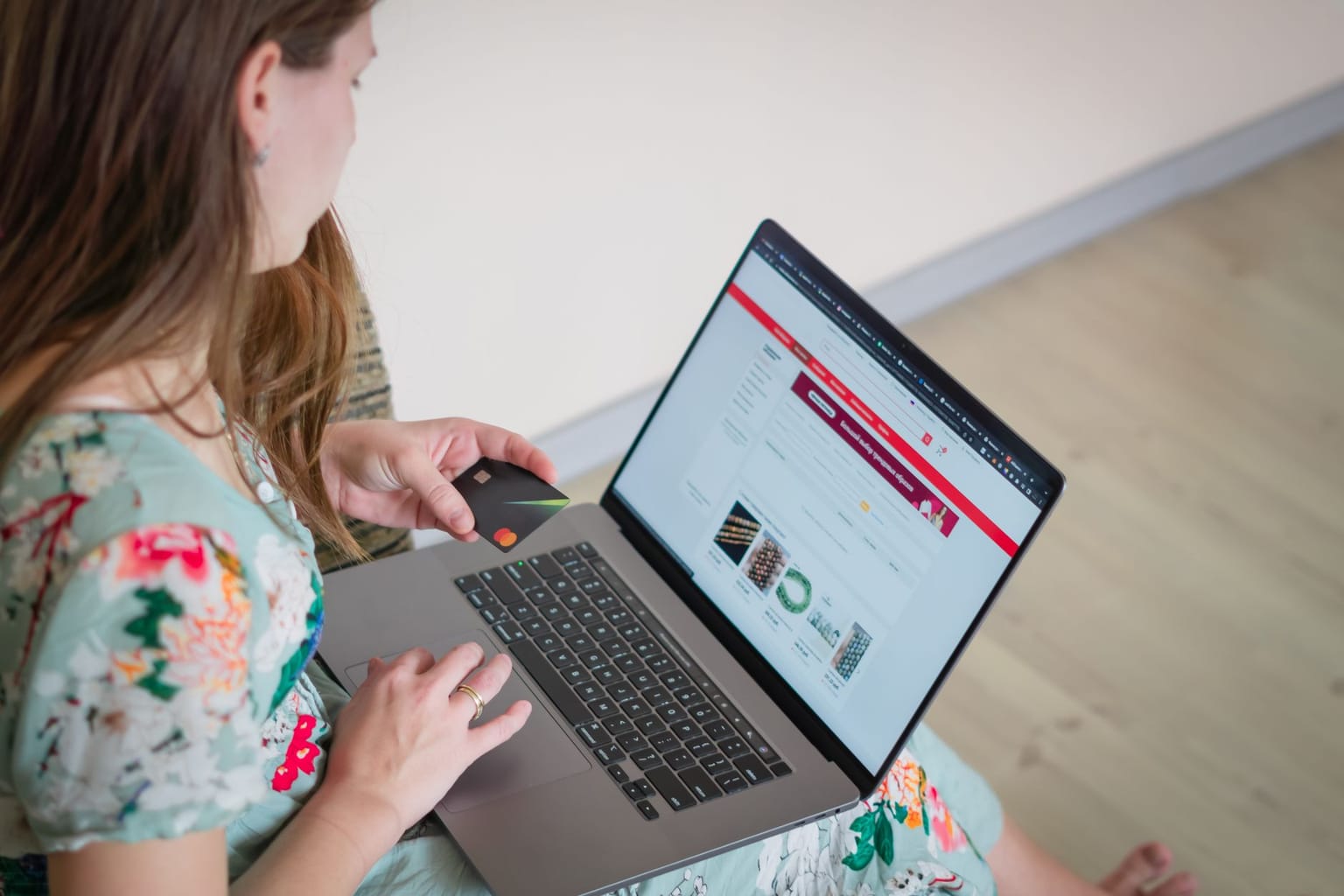 A woman in a floral dress sits on the floor holding a bank card and shopping online for software deals using her laptop