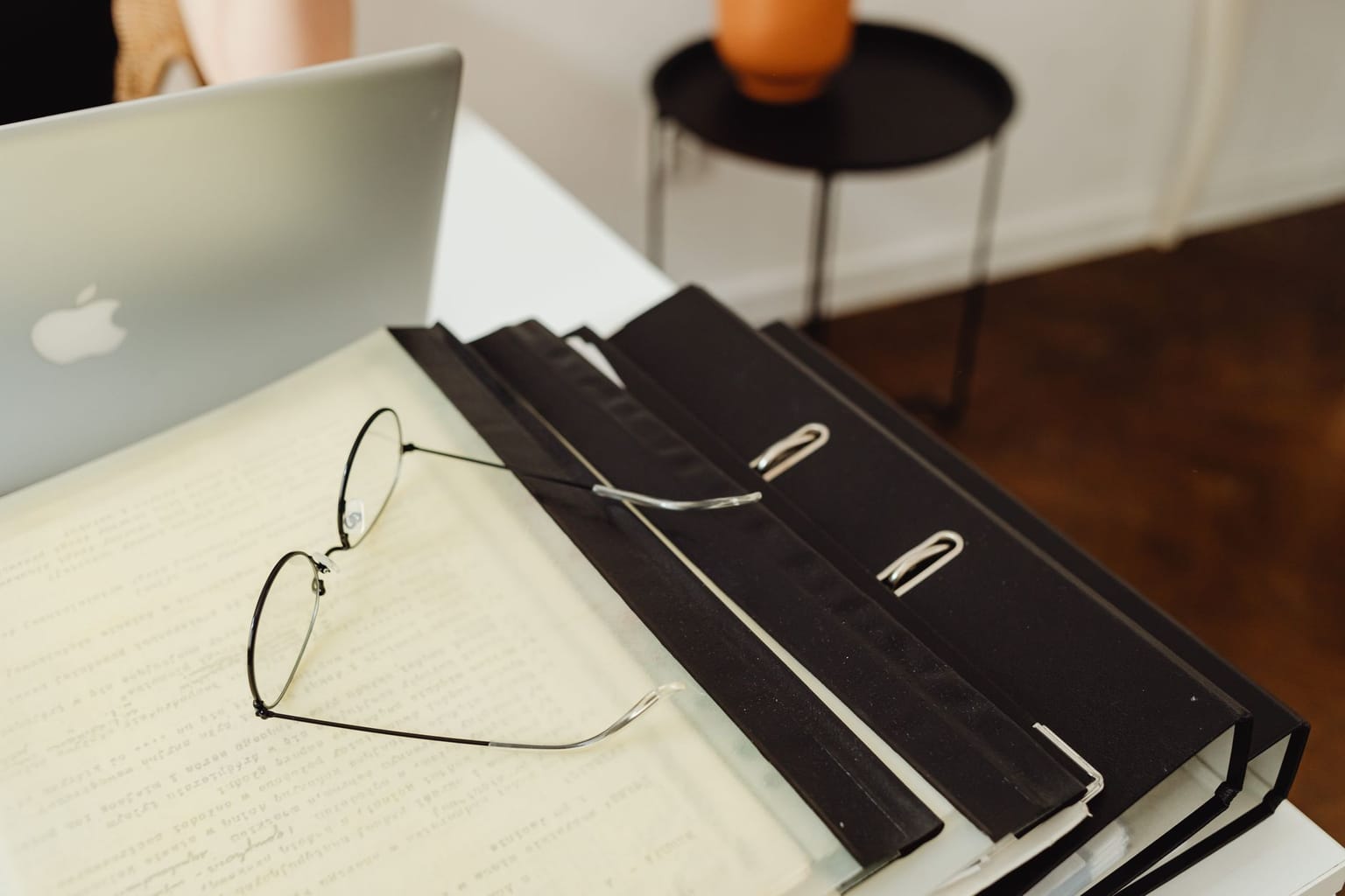 A pair of eyeglasses rests on open documents in a black binder beside a laptop on a white desk, useful for hunting lifetime software deals