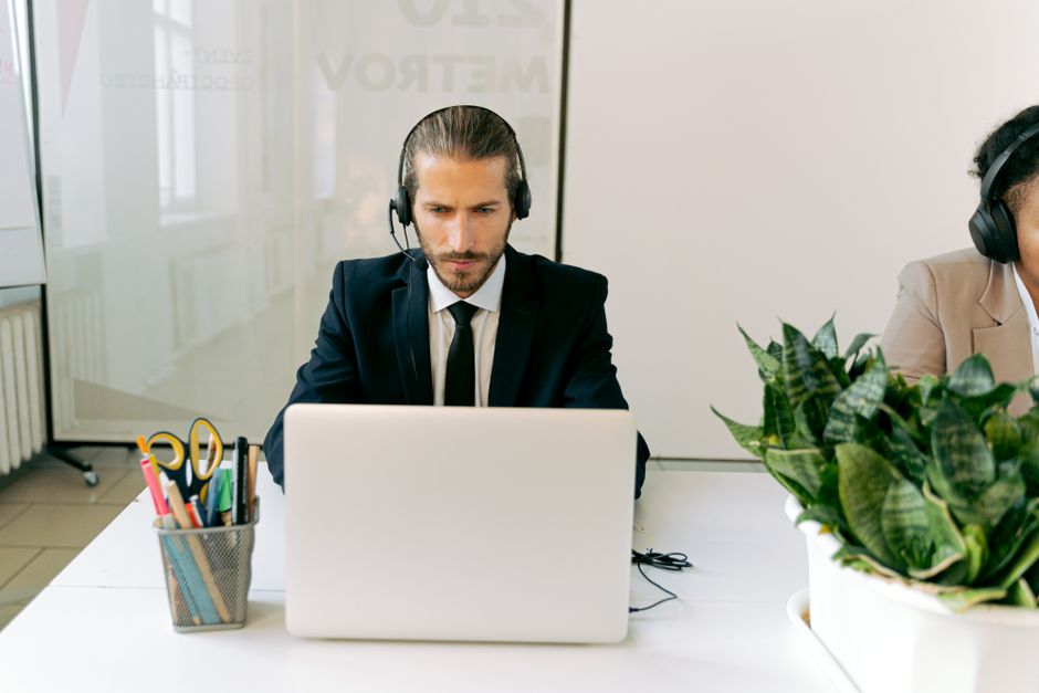 A man in a suit with a headset, using customer relationship management software, works on a laptop at a white desk with office supplies and a plant