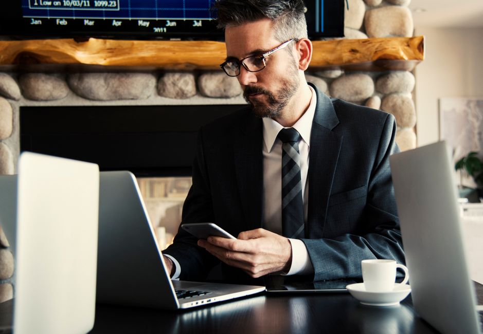A man in a suit sits at a desk with a laptop, holding a smartphone, with a cup of coffee and a second laptop beside him