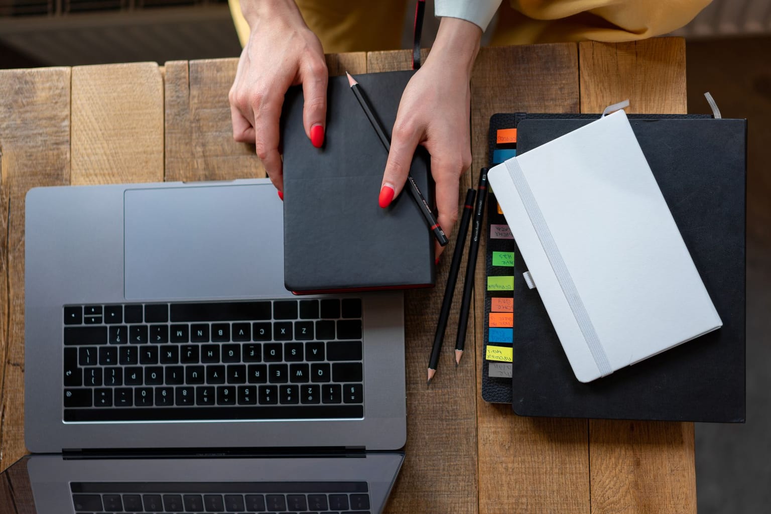 A person holds a black notebook over a laptop on a wooden table, surrounded by colored-tabbed notebooks and pencils