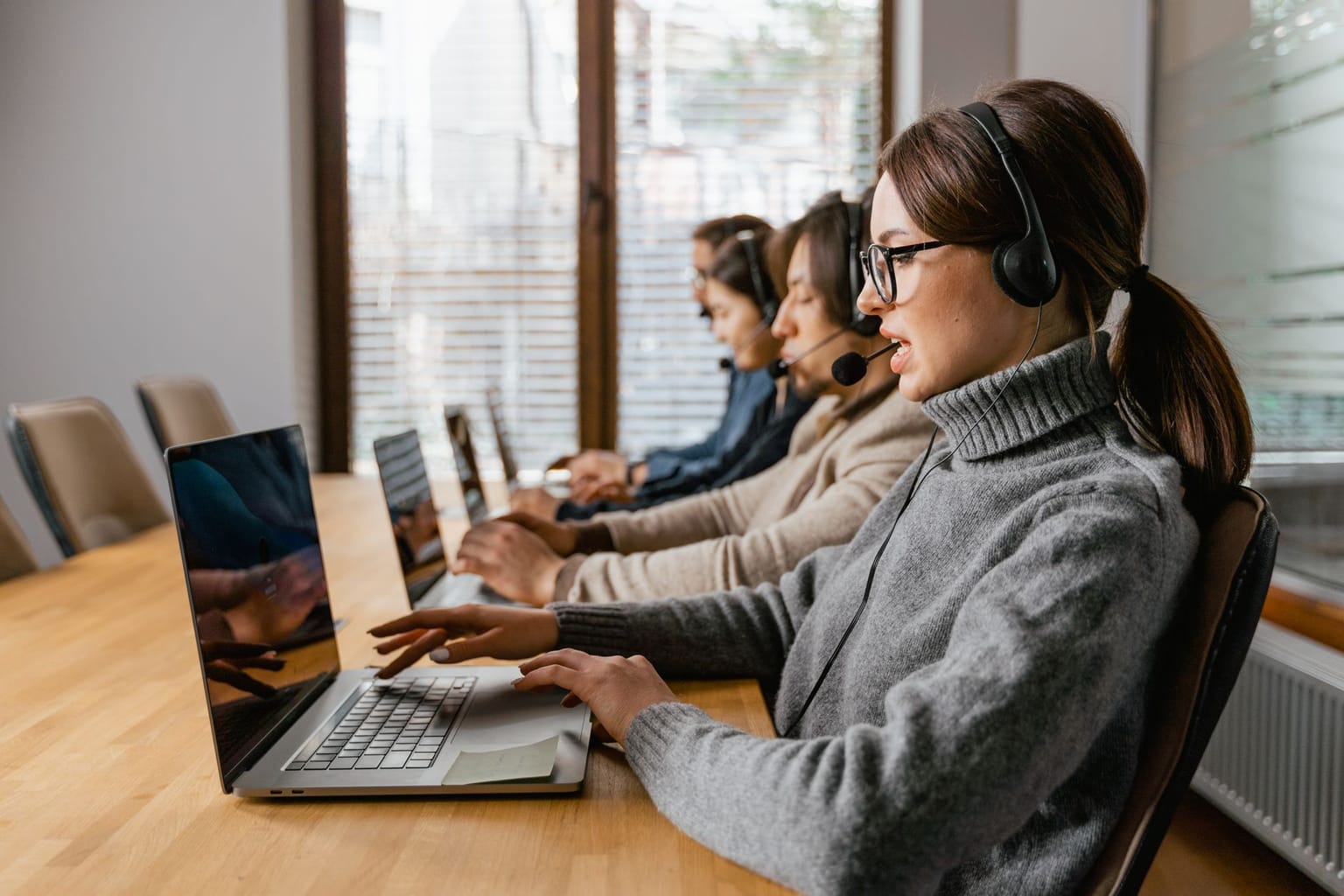Four people wearing headsets work on laptops at a wooden table in an office, providing customer support or telecommunication services with the help