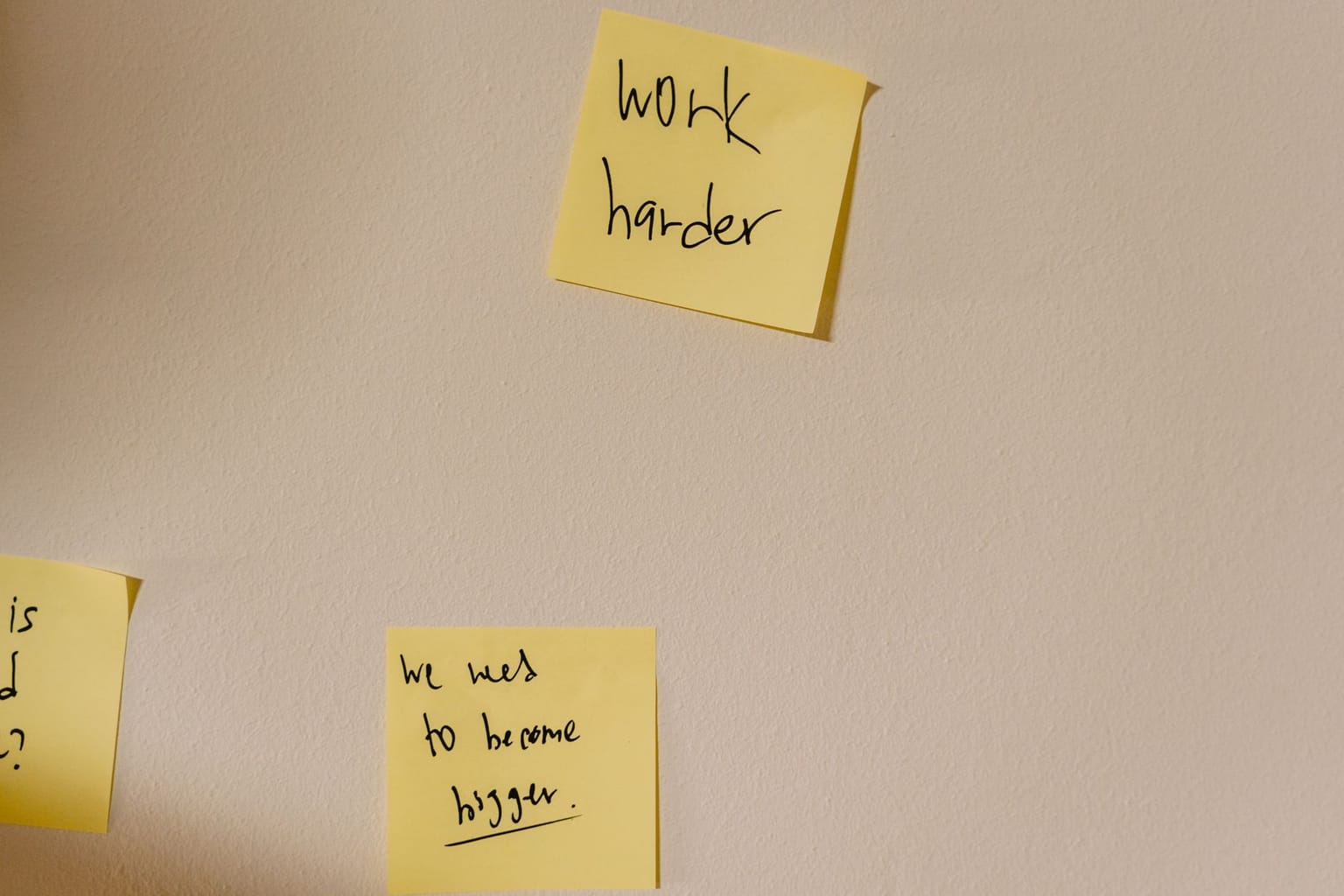 Two yellow sticky notes on a wall used as organizing tools, one reading work harder and the other we need to become bigger