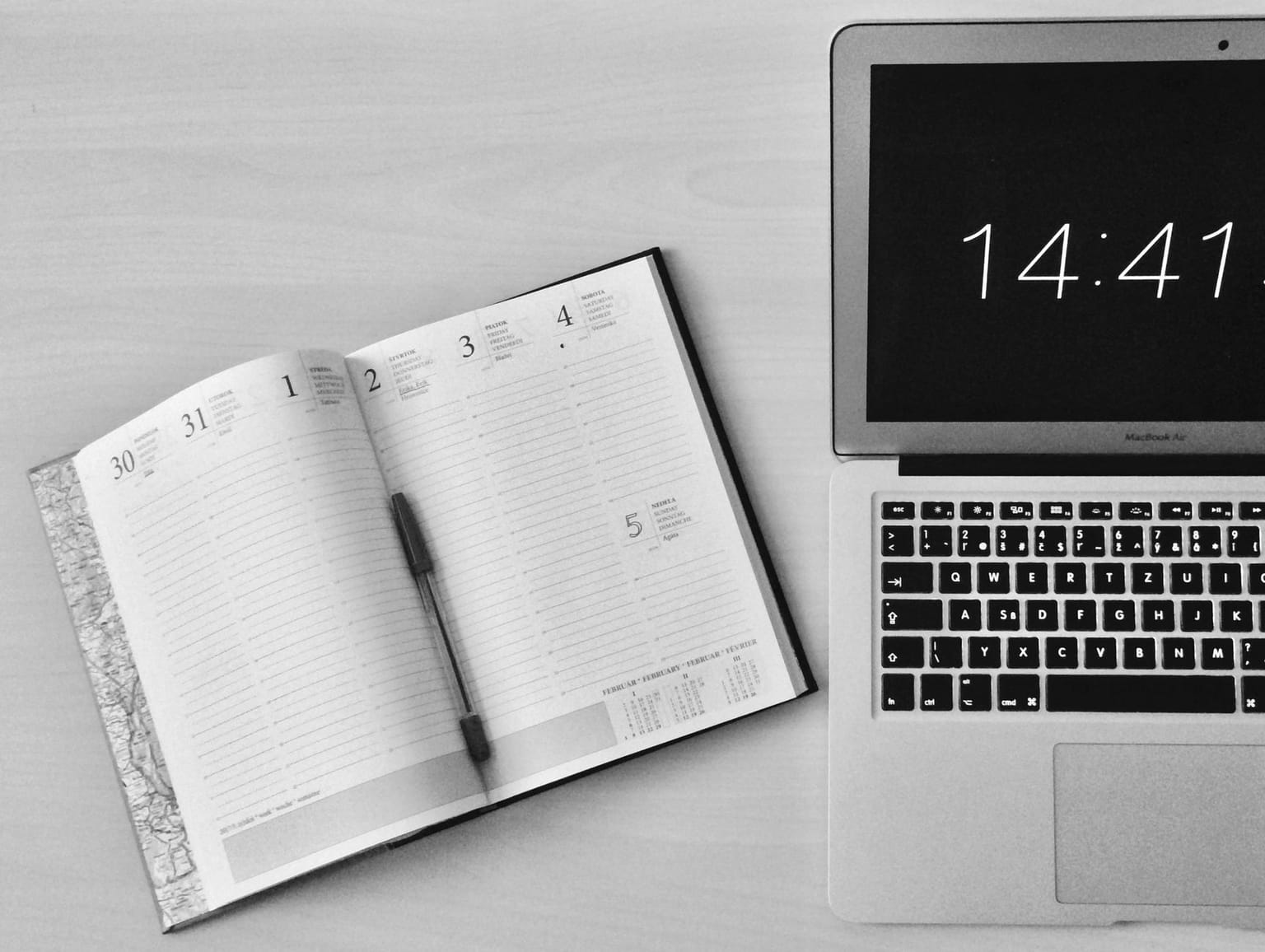 An open planner with a pen rests on a desk next to a laptop displaying the time, in black and white