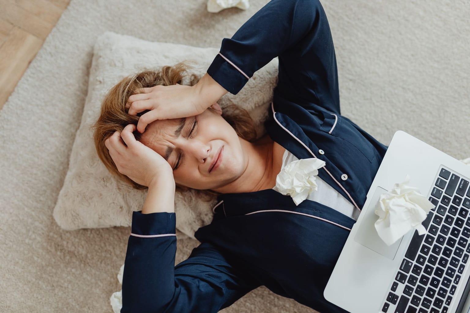 A woman in pajamas lies on a pillow on the floor, holding her head in frustration as she sits next to a laptop