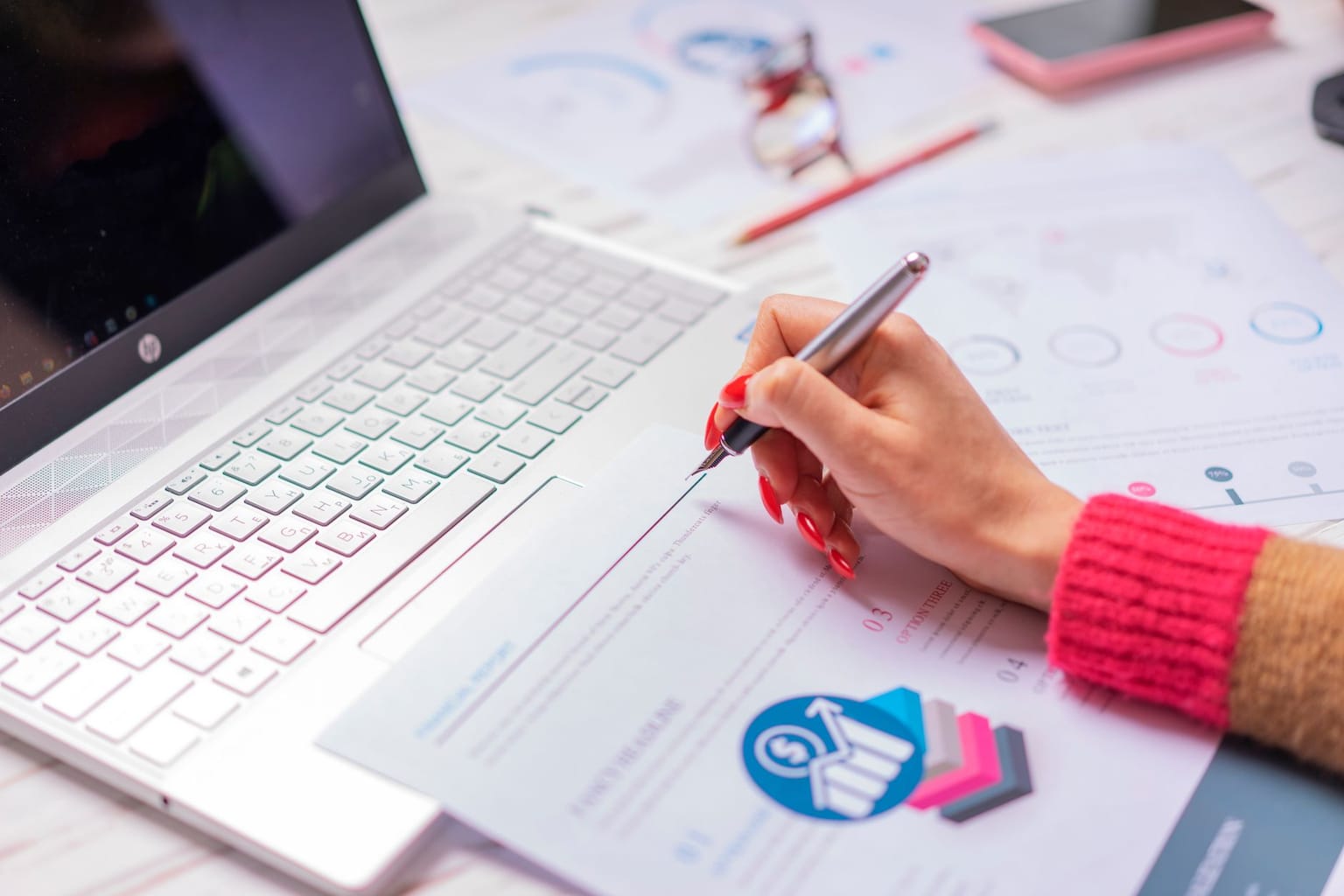 A person with red nails is holding a pen and reviewing printed charts and graphs next to an open laptop on a desk, using an LTD tracker to review spending