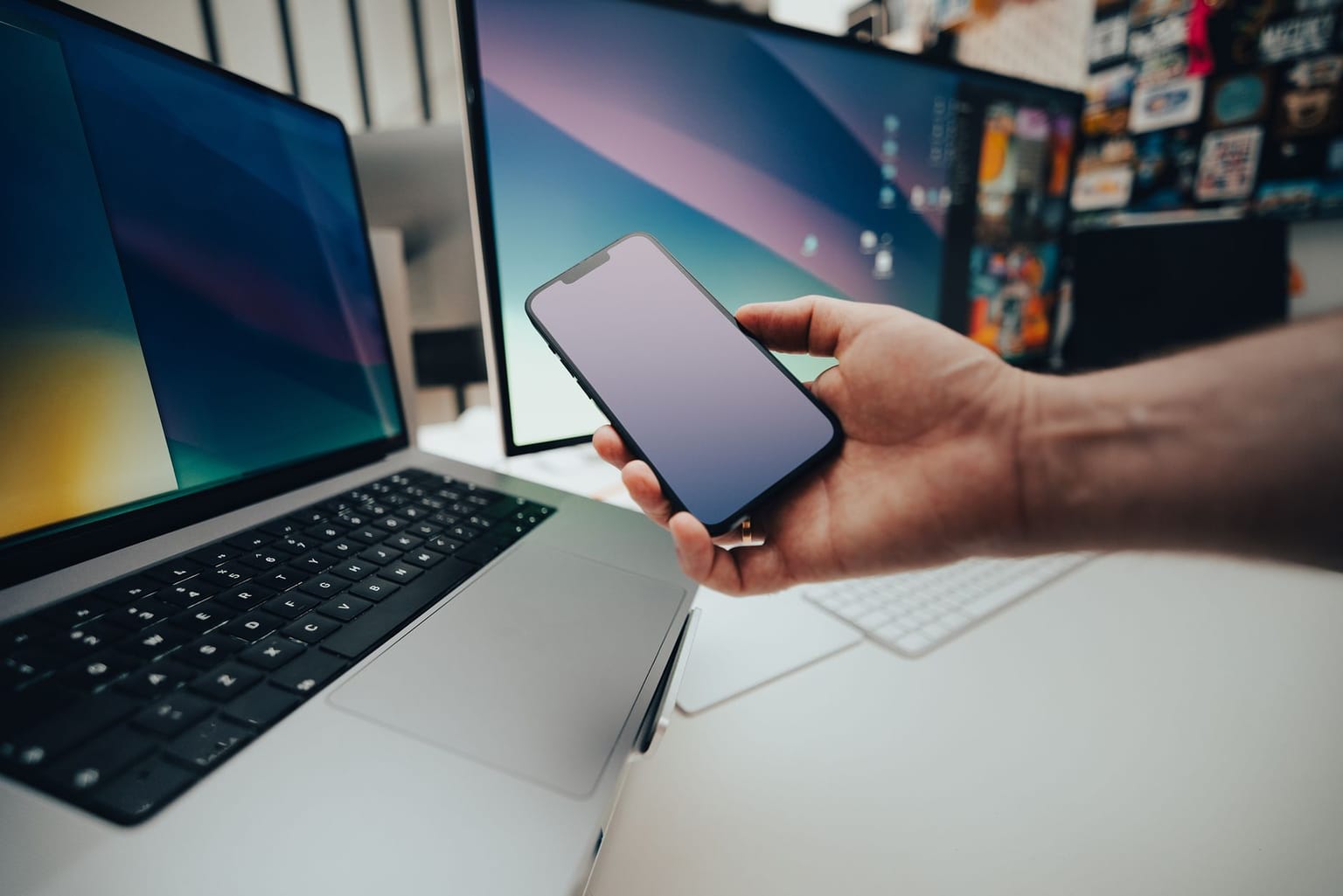 A hand holds a smartphone in front of a laptop and two computer monitors on a white desk in a modern workspace