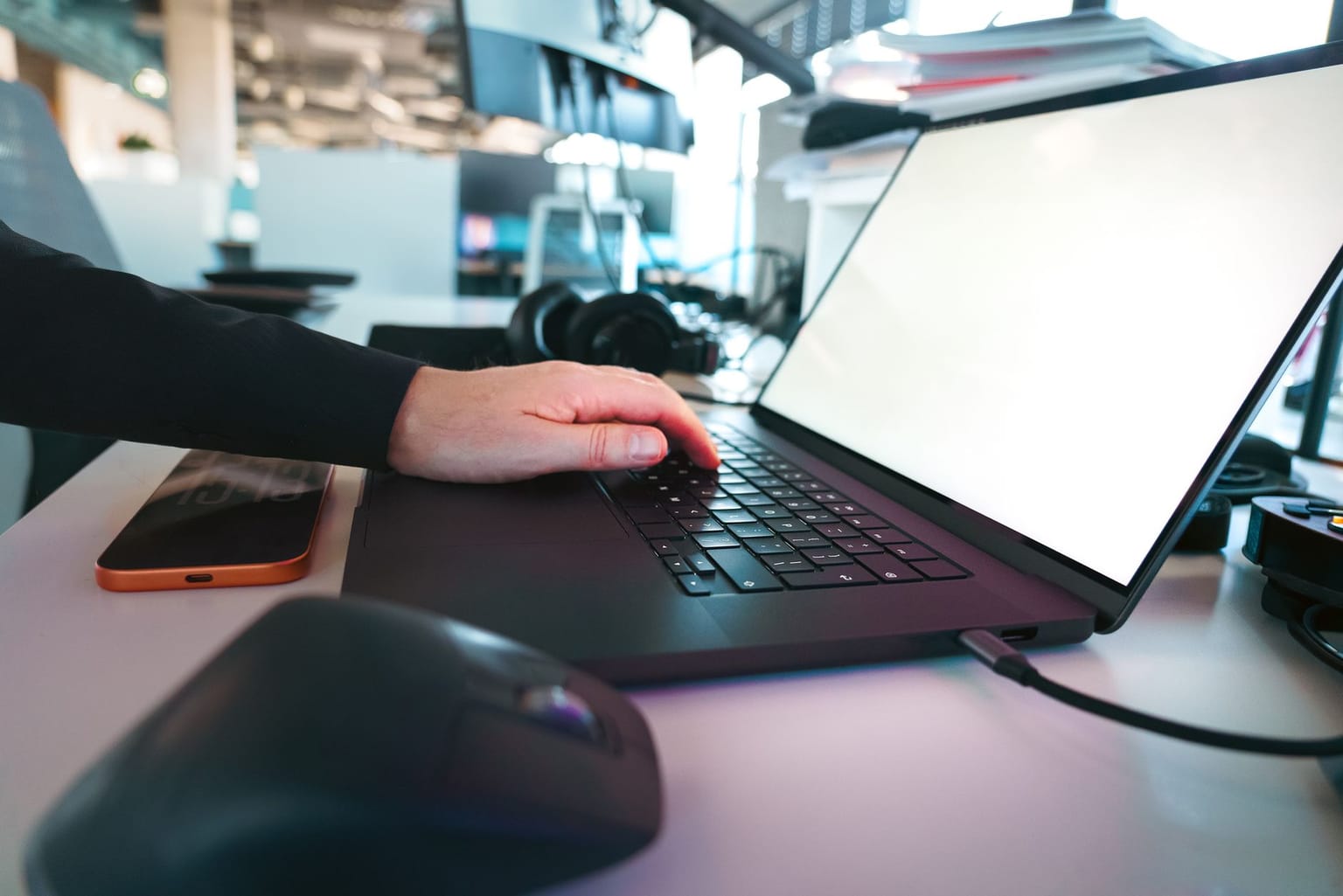 A person’s hand types on a black laptop keyboard at a desk with a computer mouse, smartphone, and headphones nearby in a modern office setting