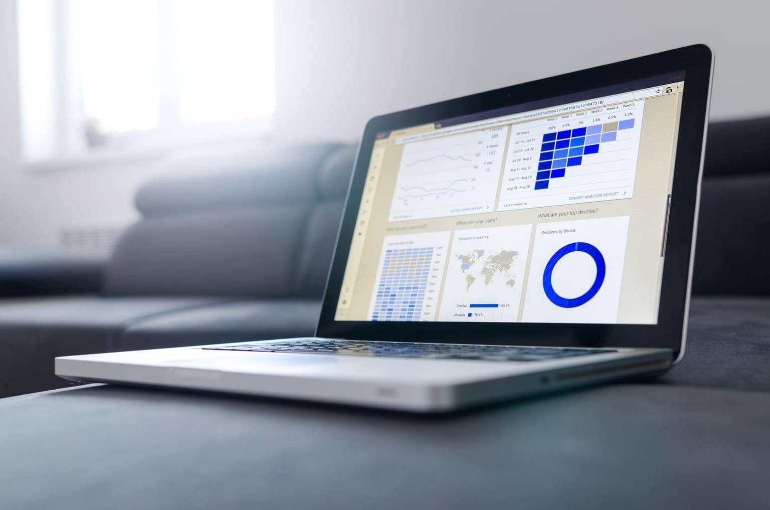 A laptop sits on a gray couch, displaying data visualizations including charts, graphs, a world map, and a pie chart on its screen in a bright