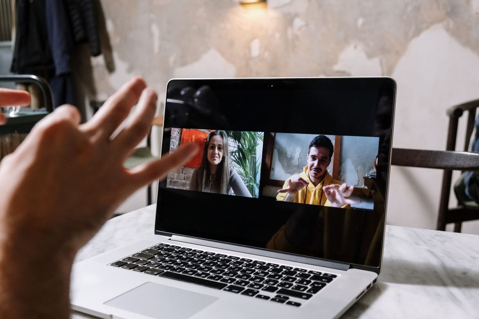 A person waves at a laptop screen showing a video call with two smiling people gesturing in a cozy indoor setting