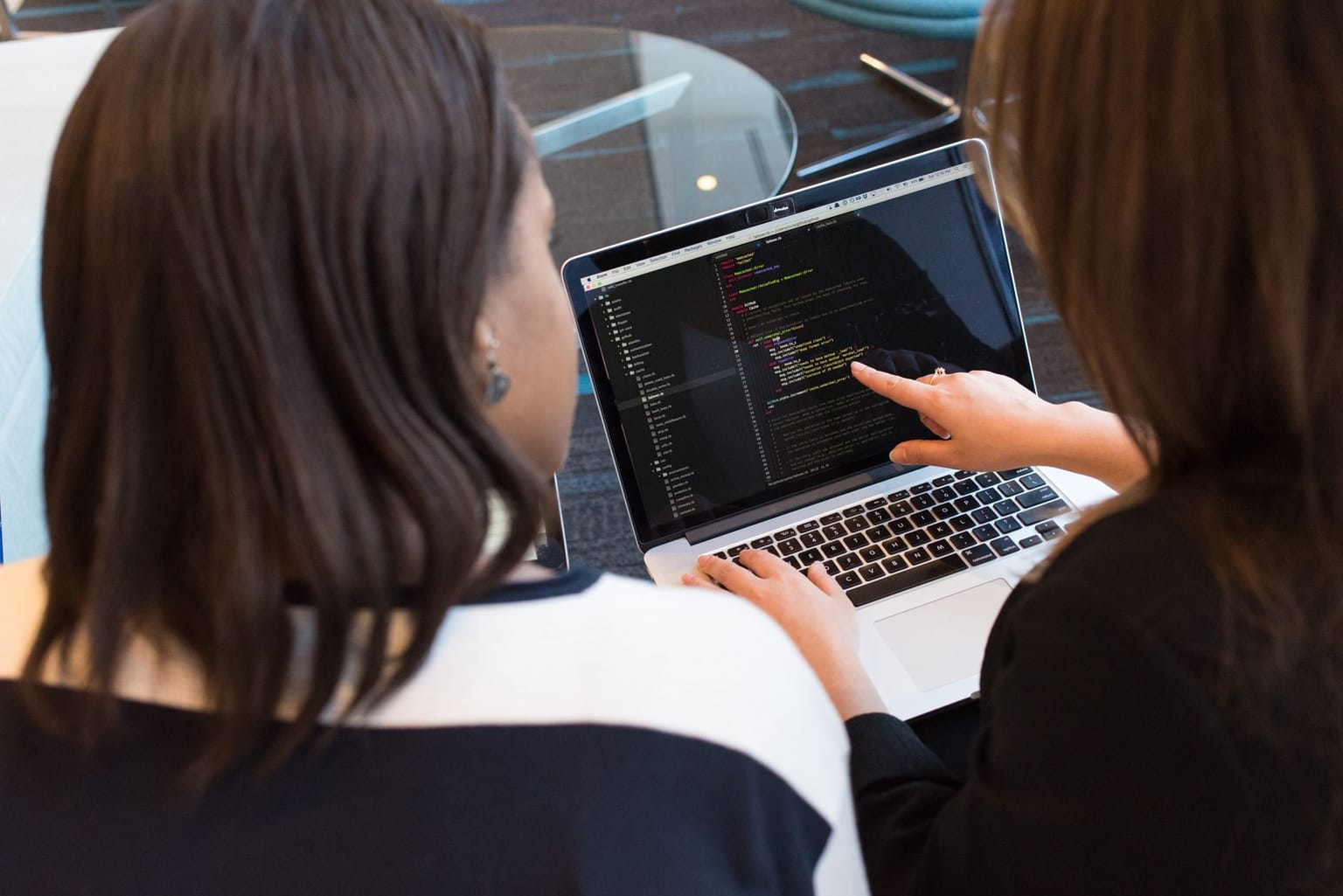 Two people sit together looking at a laptop screen displaying lines of code; one person is pointing at the code while the other observes
