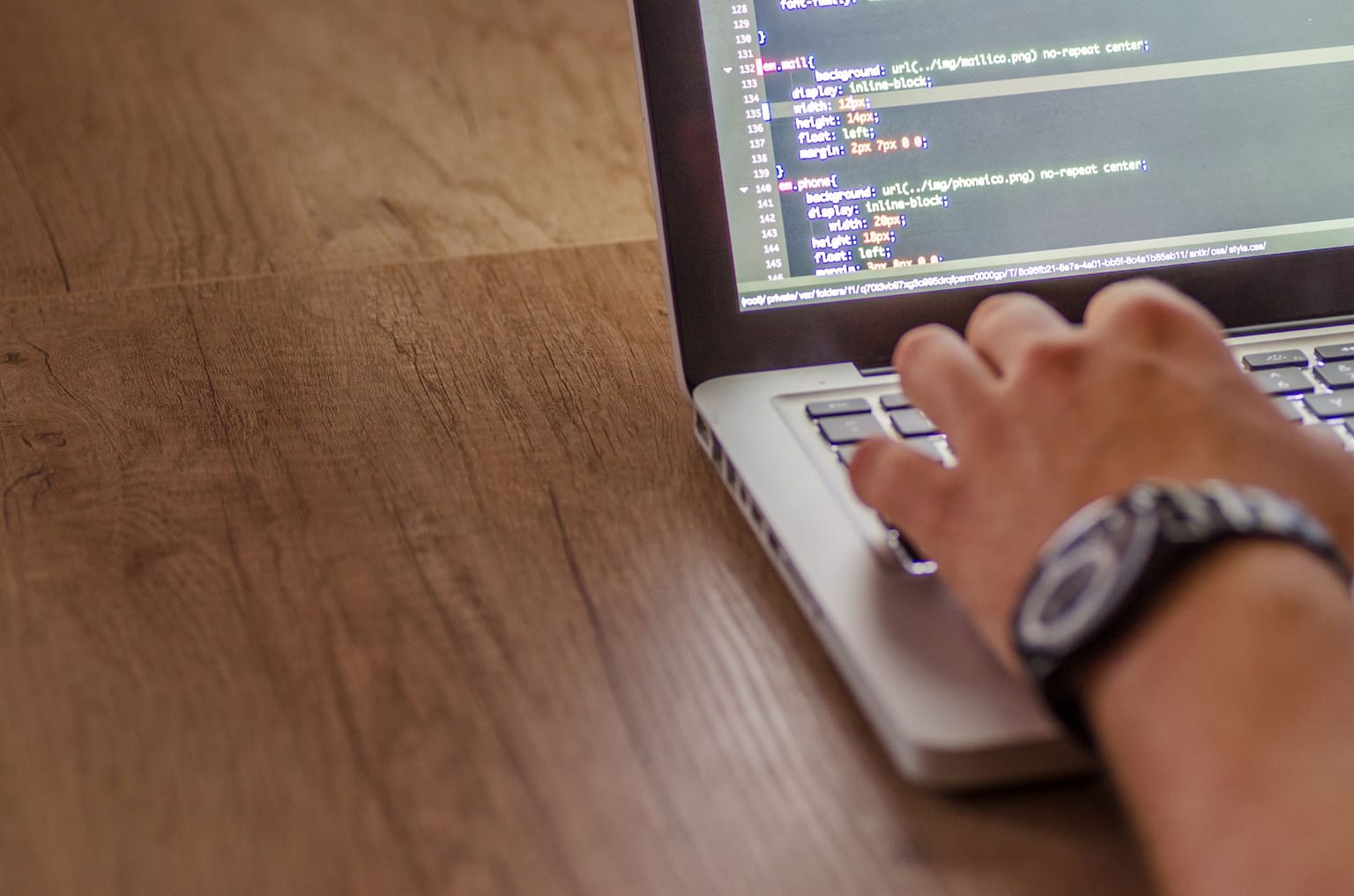 A person wearing a watch types on a laptop displaying colorful lines of code on the screen, sitting at a wooden desk