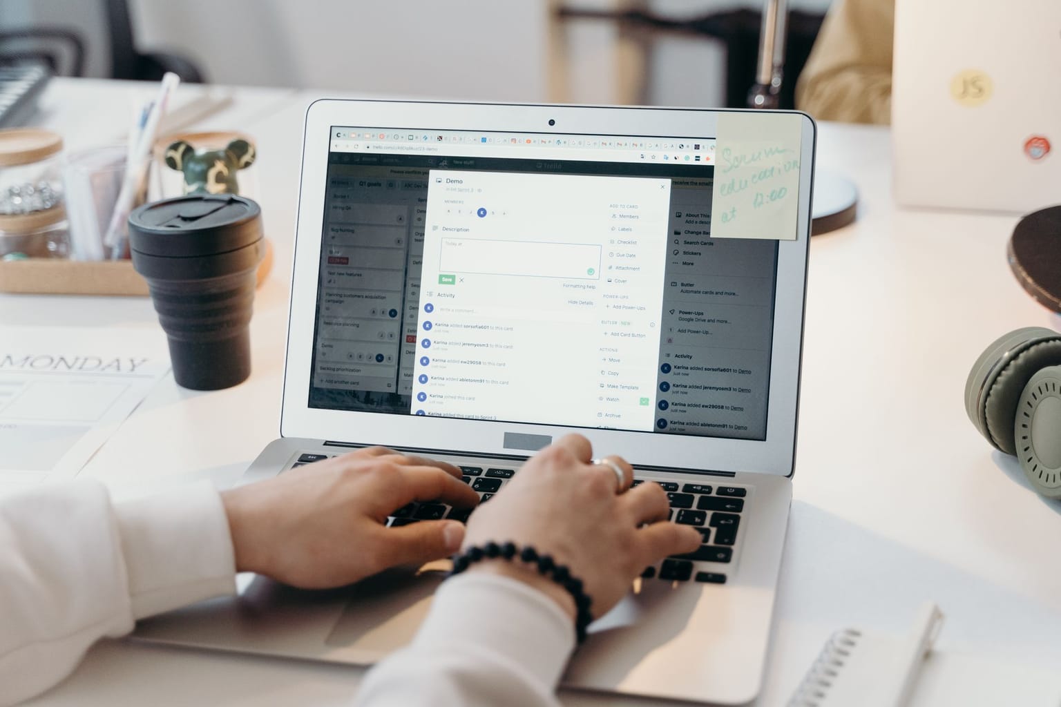 A person types on a laptop at a desk with a coffee cup, headphones, and a sticky note on the laptop. The laptop screen displays an email marketing tool