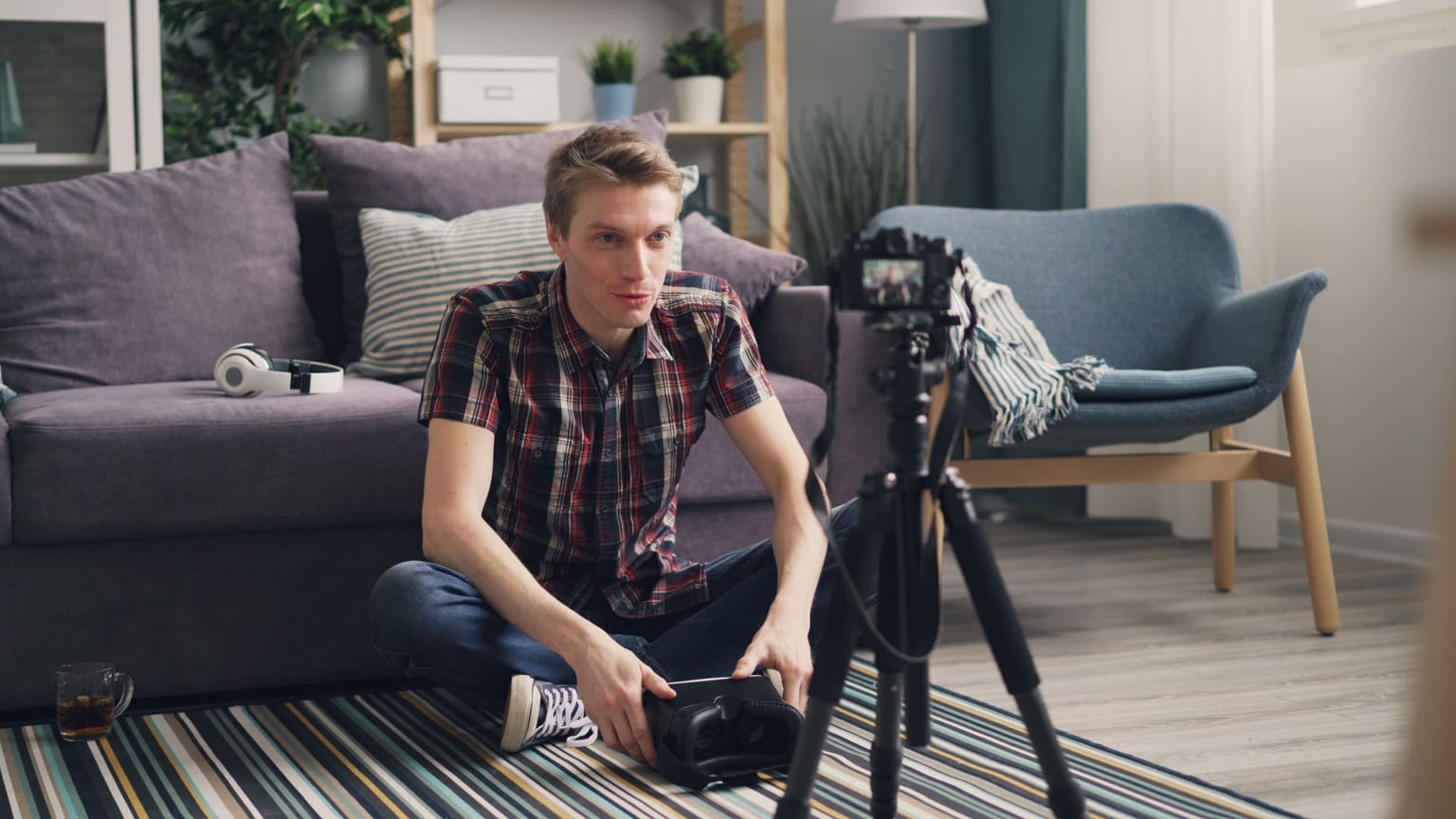 A content creator sits cross-legged on the floor, facing a camera set on a tripod. There’s a sofa, headphones, and a glass nearby in a cozy