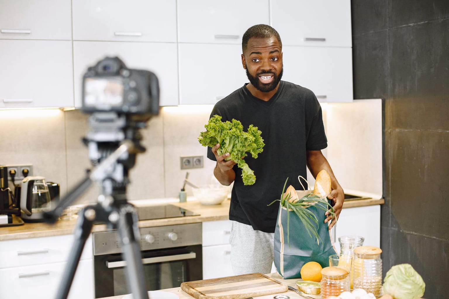 A man stands in a modern kitchen holding lettuce and a grocery bag, smiling at a camera on a tripod—clearly comfortable in his role as a content creator