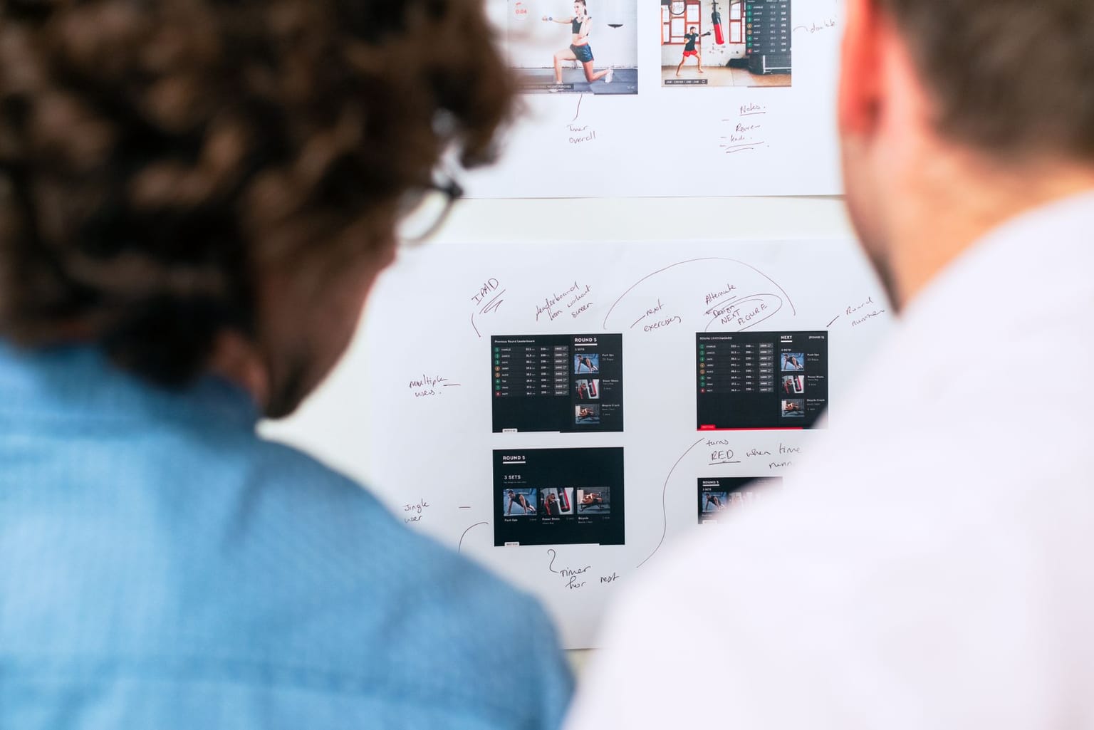Two people look at a whiteboard with printed website or app screenshots and handwritten notes
