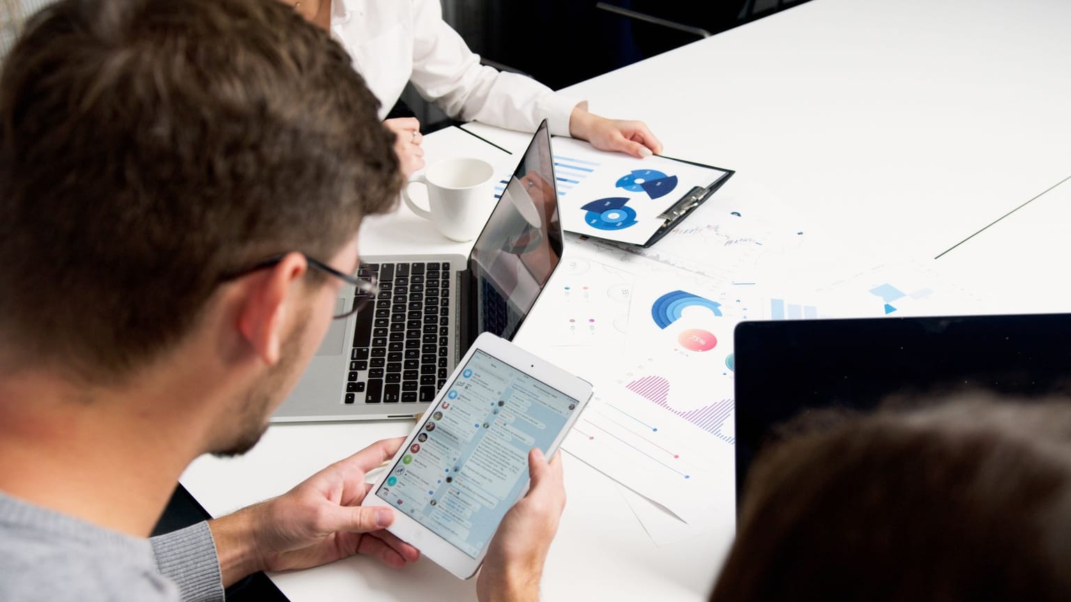 Three people sit at a table using a tablet, laptop, and a clipboard with charts. Papers with graphs and a coffee cup are also on the white table