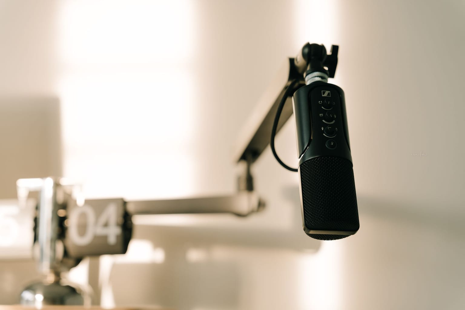 A black microphone on an adjustable arm stand, ideal for podcast recording, is positioned near a blurred wall clock showing “04” in the background