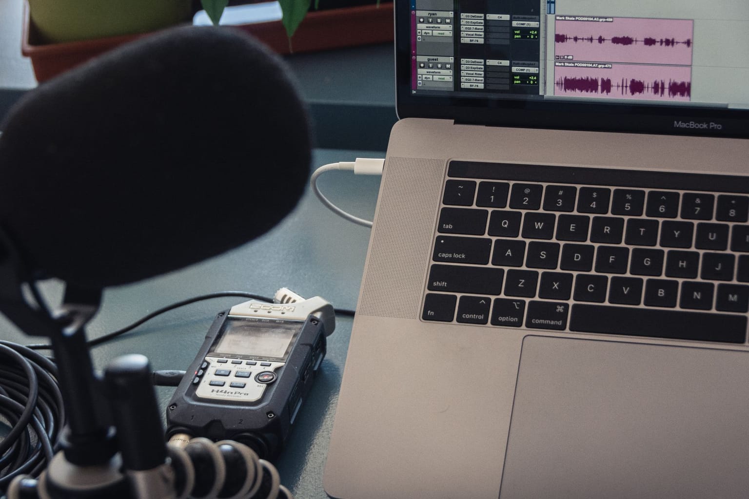 A close-up of a laptop with audio editing software open, a microphone, and a portable audio recorder on a desk