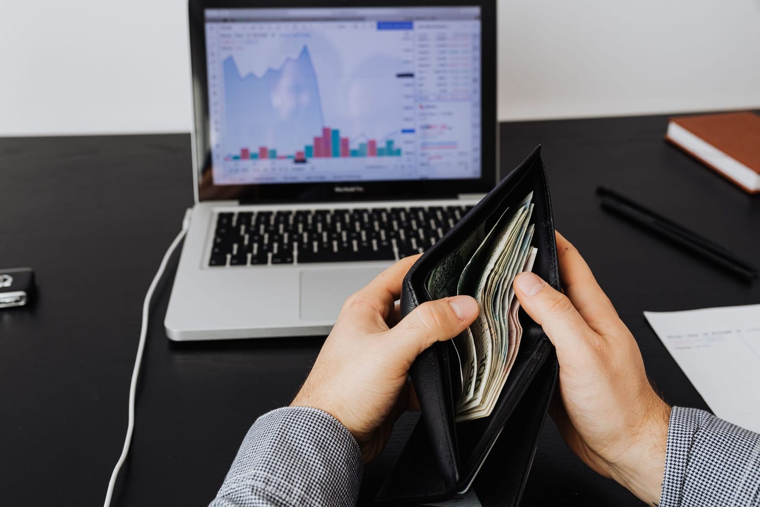 A person holding an open wallet with cash in front of a laptop displaying a stock market chart, on a desk with a pen, notebook