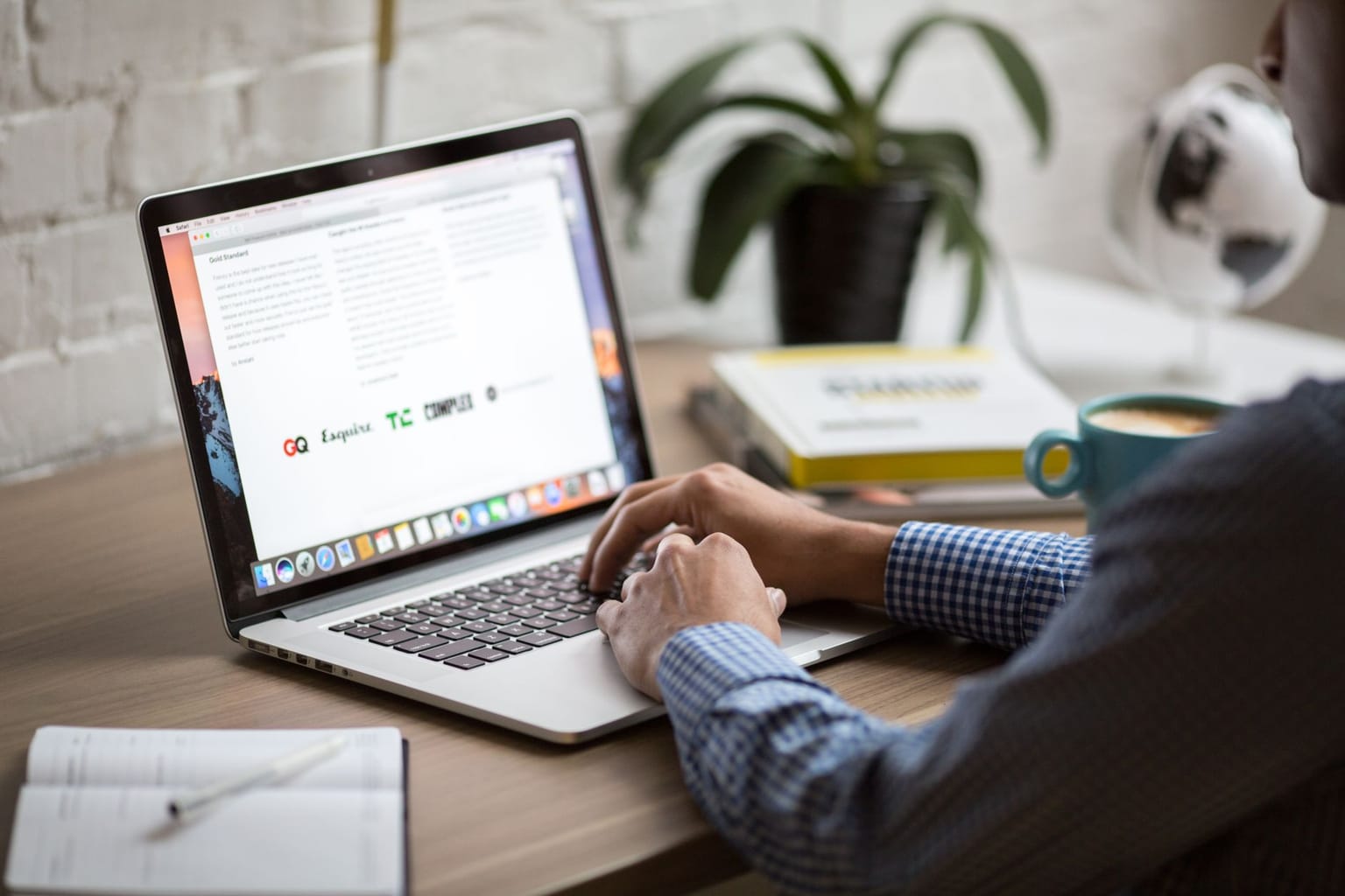 A person types on a laptop at a wooden desk, planning an email marketing campaign. A notebook, cup of coffee, potted plant, stack of books