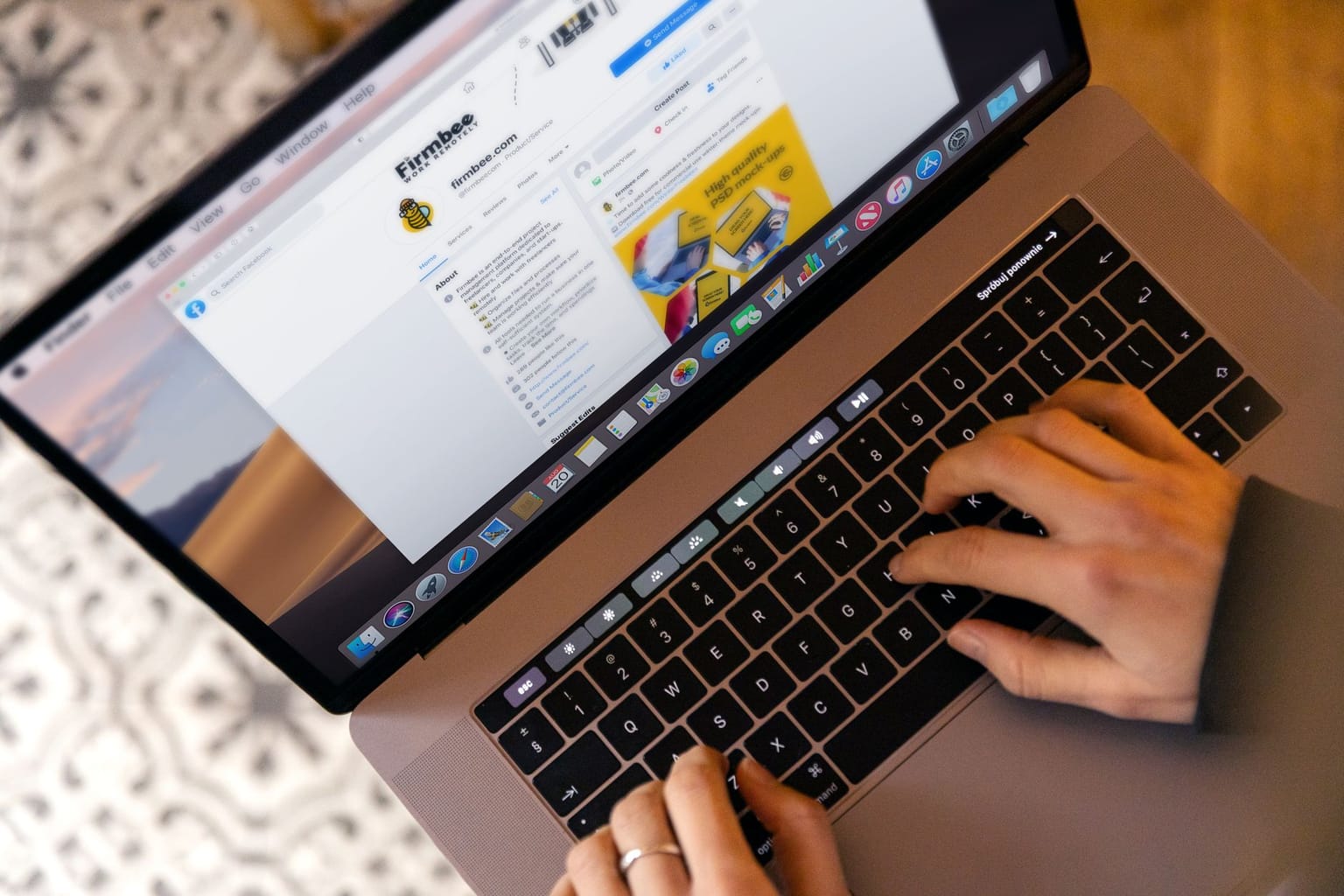 A person types on a MacBook laptop displaying a Facebook page, with both hands on the keyboard. The laptop is on a patterned surface