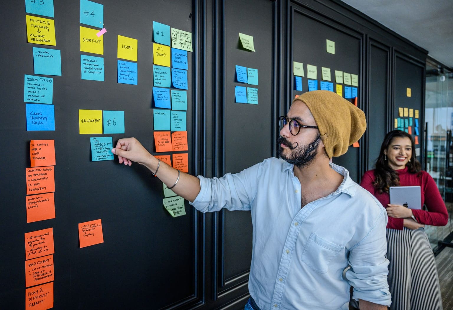 A man in a beanie places sticky notes on a black wall covered with colorful notes, illustrating an engaging project management session