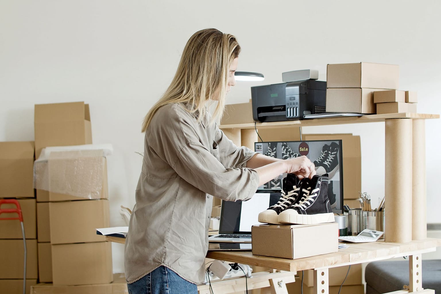A woman packs black-and-white sneakers into a cardboard box on a desk surrounded by shipping boxes, a computer