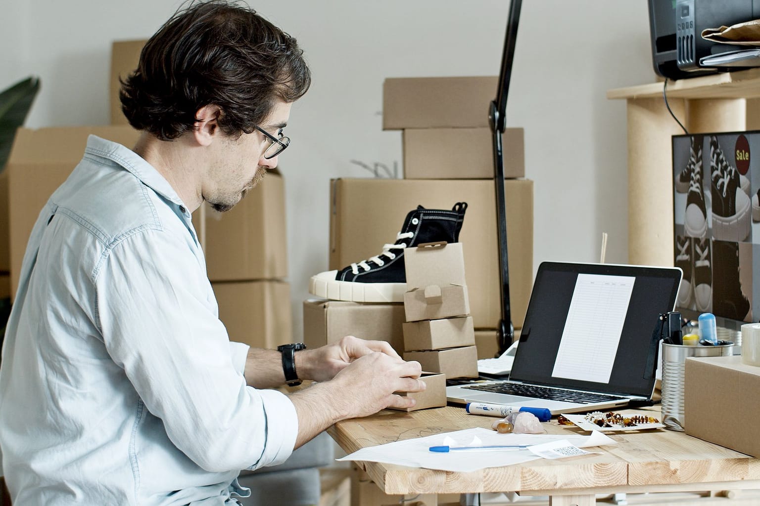 A man sits at a desk assembling a cardboard box, with a laptop displaying WooCommerce product options, sneakers, small boxes