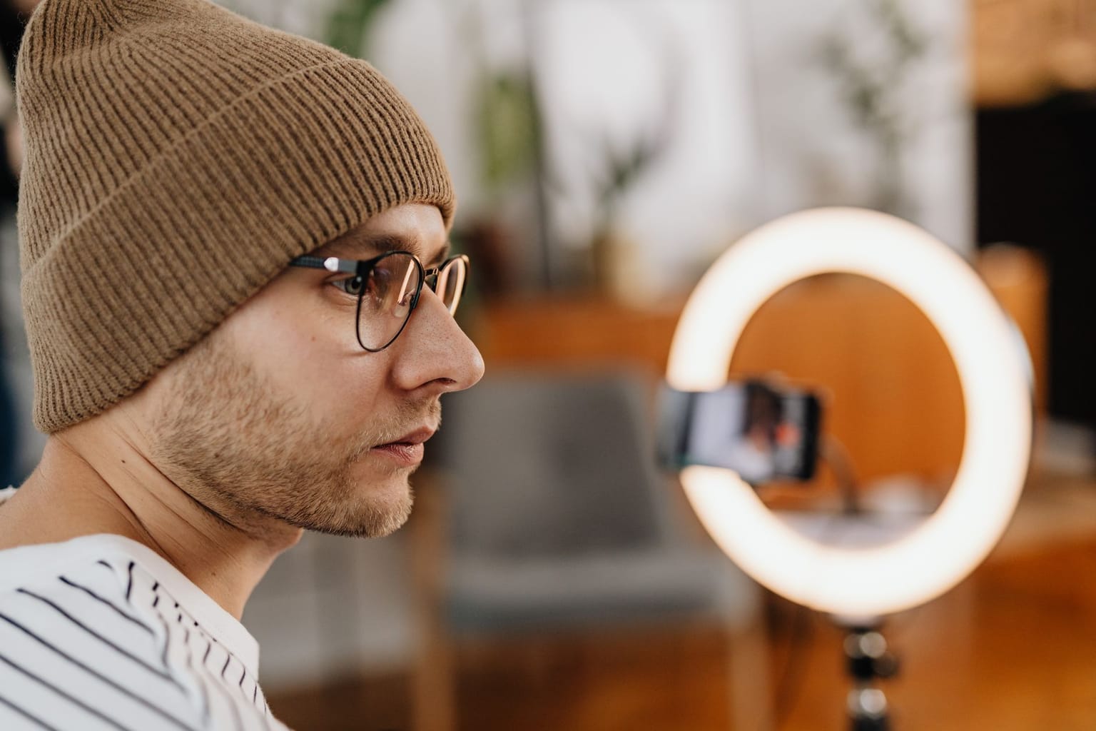 A man wearing glasses and a brown beanie sits in front of a ring light and smartphone