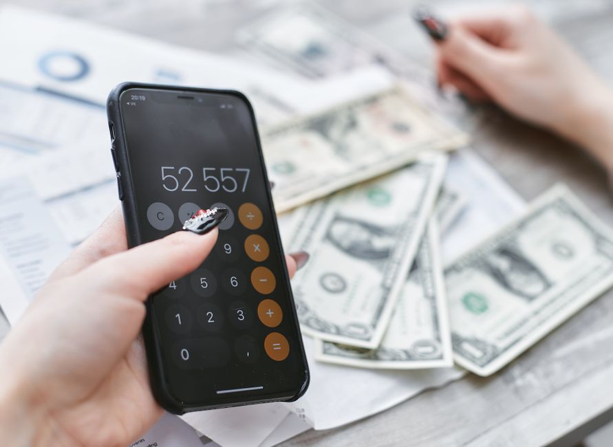 A person uses a calculator app on a smartphone while U.S. dollar bills and financial papers are spread on a table.